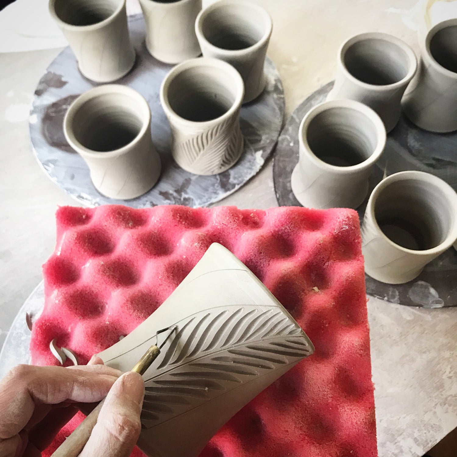 Joy tanner cutting into one of her in process cups in the studio on a red piece of foam, other in process cups in the background.