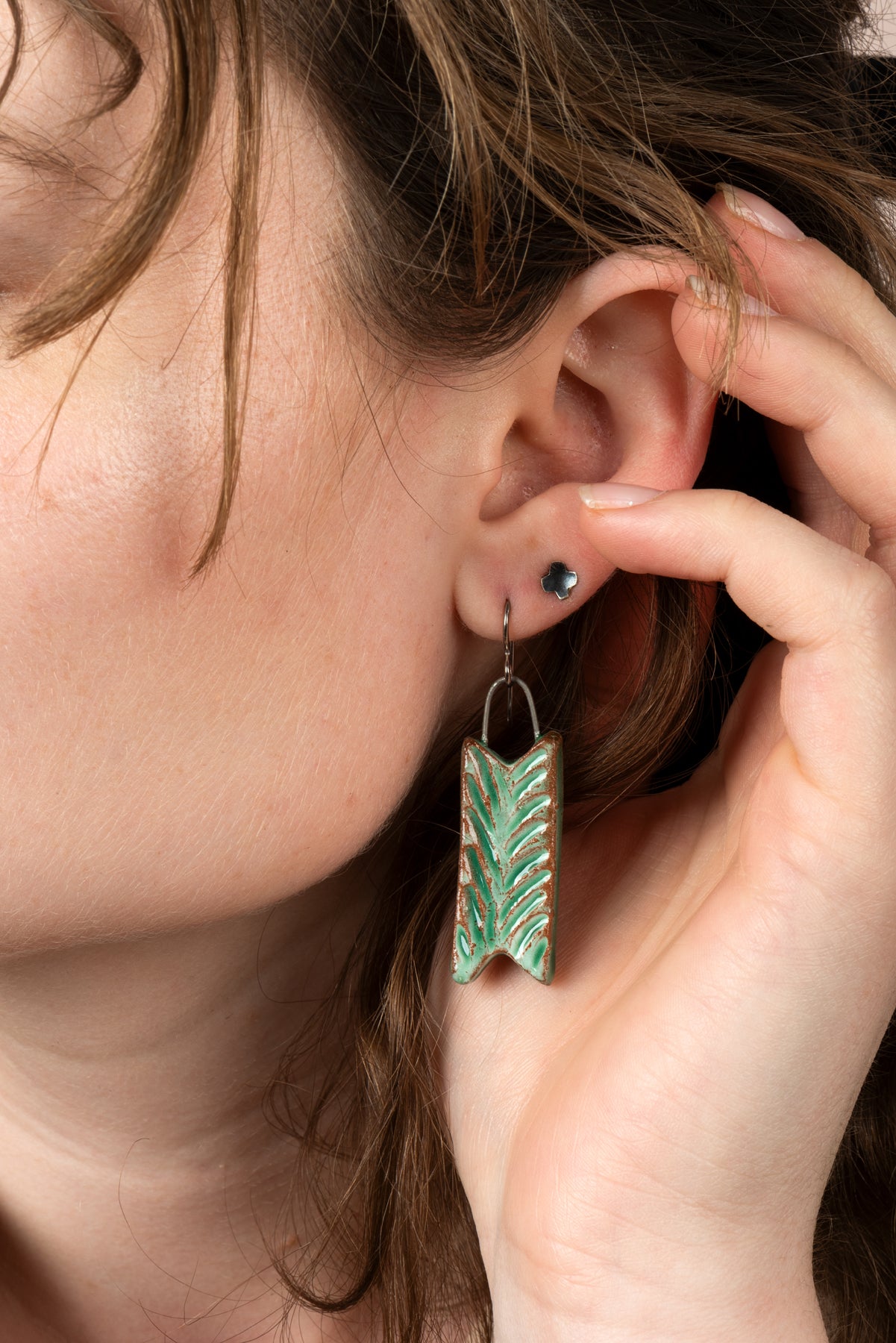 A closeup of one of Joy Tanner's green carved ceramic earrings hanging in someone's ear, their hand reaching up to brush their hair back. Image by Loam