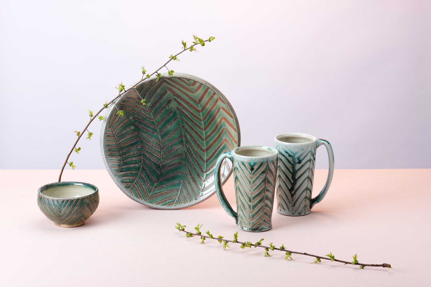 A group of turquoise pottery, handmade by artist Joy Tanner, sits on a pink tabletop against a purpleish backdrop. New Spring twigs lean on a propped up plate. Image by Loam.