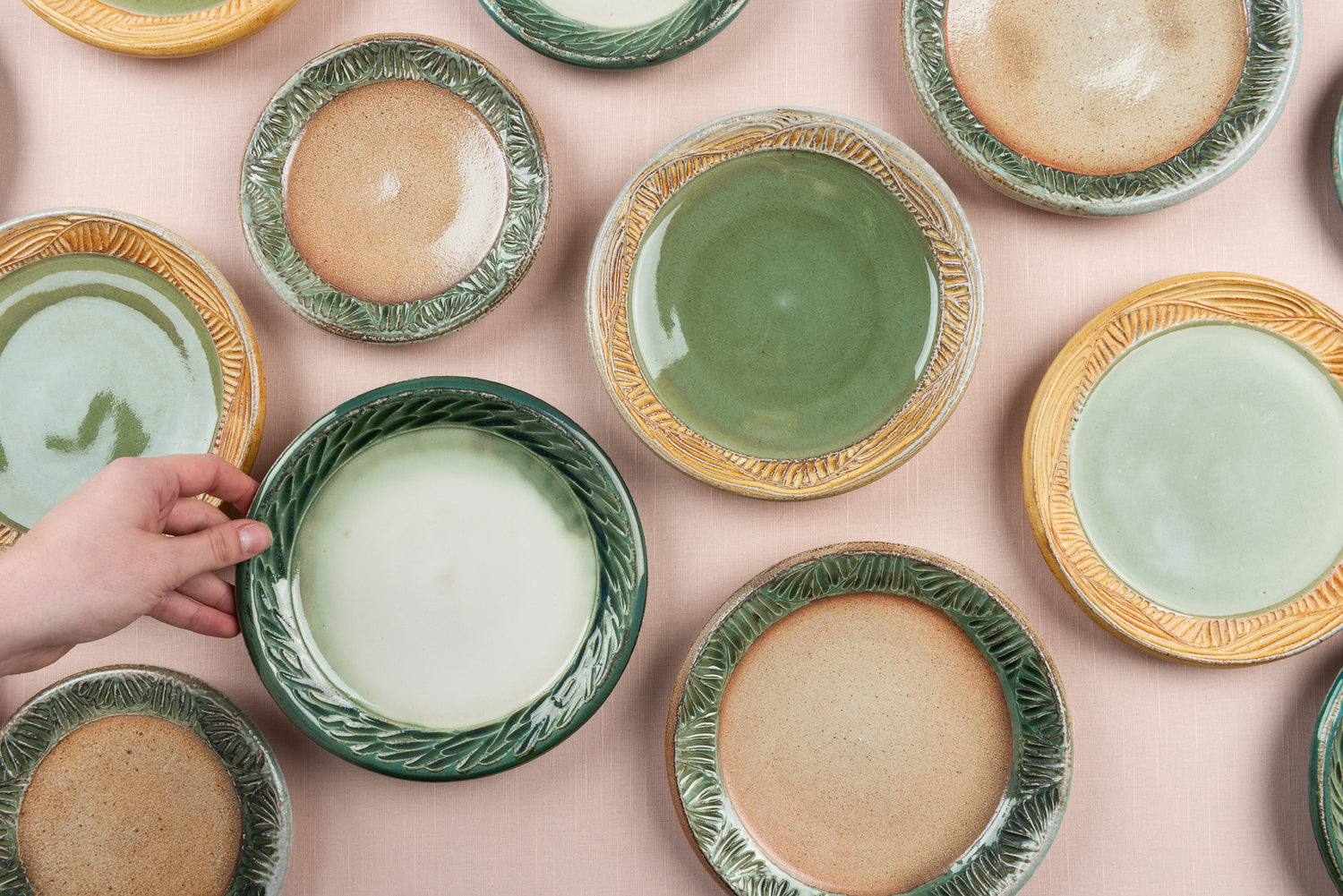Hand thrown plates in various shades of green and cream, shot from above, sit on a pink table top, a hand reaching in from out of frame to pick one up. Artist Joy Tanner.