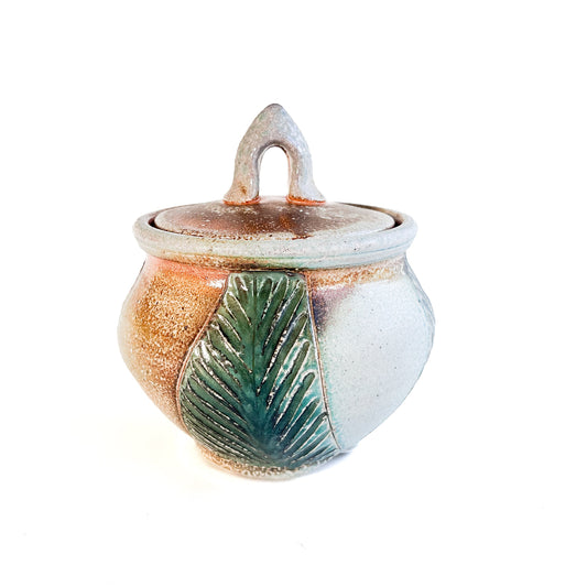 This small round handmade jar could be used to store tea bags, sugar, or candy. The green leaf pattern that is hand carved is inspired by nature.