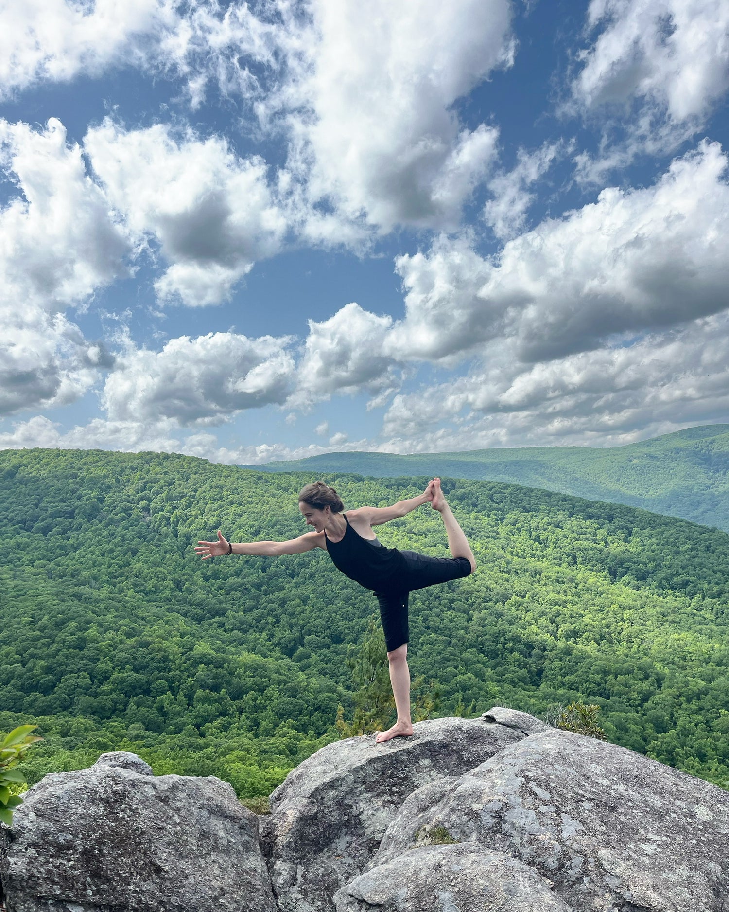 Artist at Yogi Joy tanner stands in a yoga pose on a rock against a backdrop of green rolling mountains and a blue sky with clouds . 