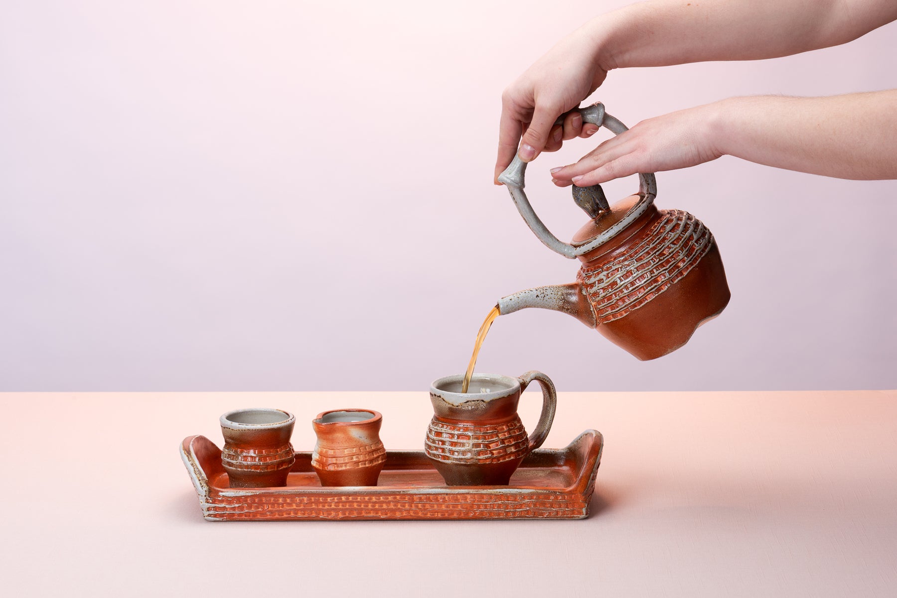 Hands come in from out of frame to pour tea out of a rusty colored Joy Tanner Pottery teapot into a little cup that sits on a handmade ceramic tray and a little creamer and sugar that sit on top of the tray as well.  
