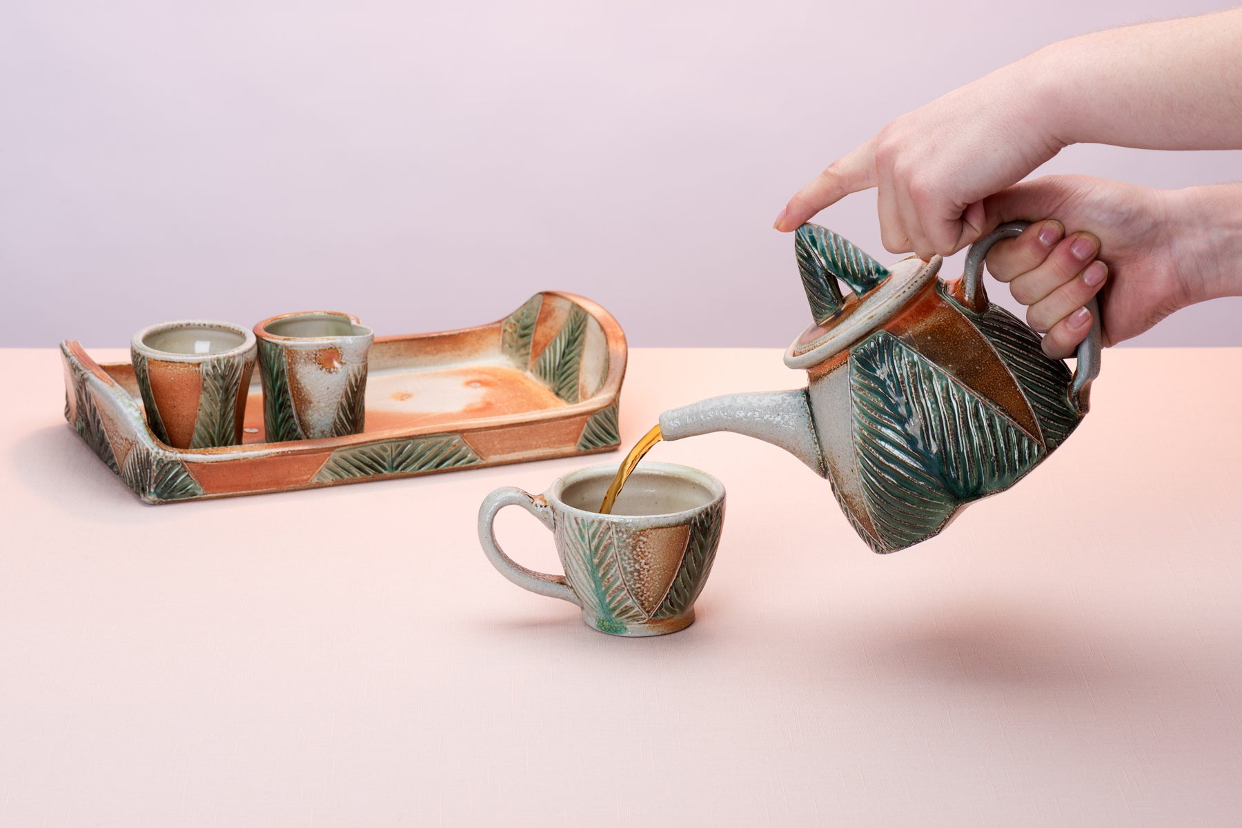 Hands come in from out of frame to pour tea out of a Joy Tanner Pottery teapot into a little cup that sits on a pink table in front of a handmade ceramic tray and a little creamer and sugar that sit on top of the tray.