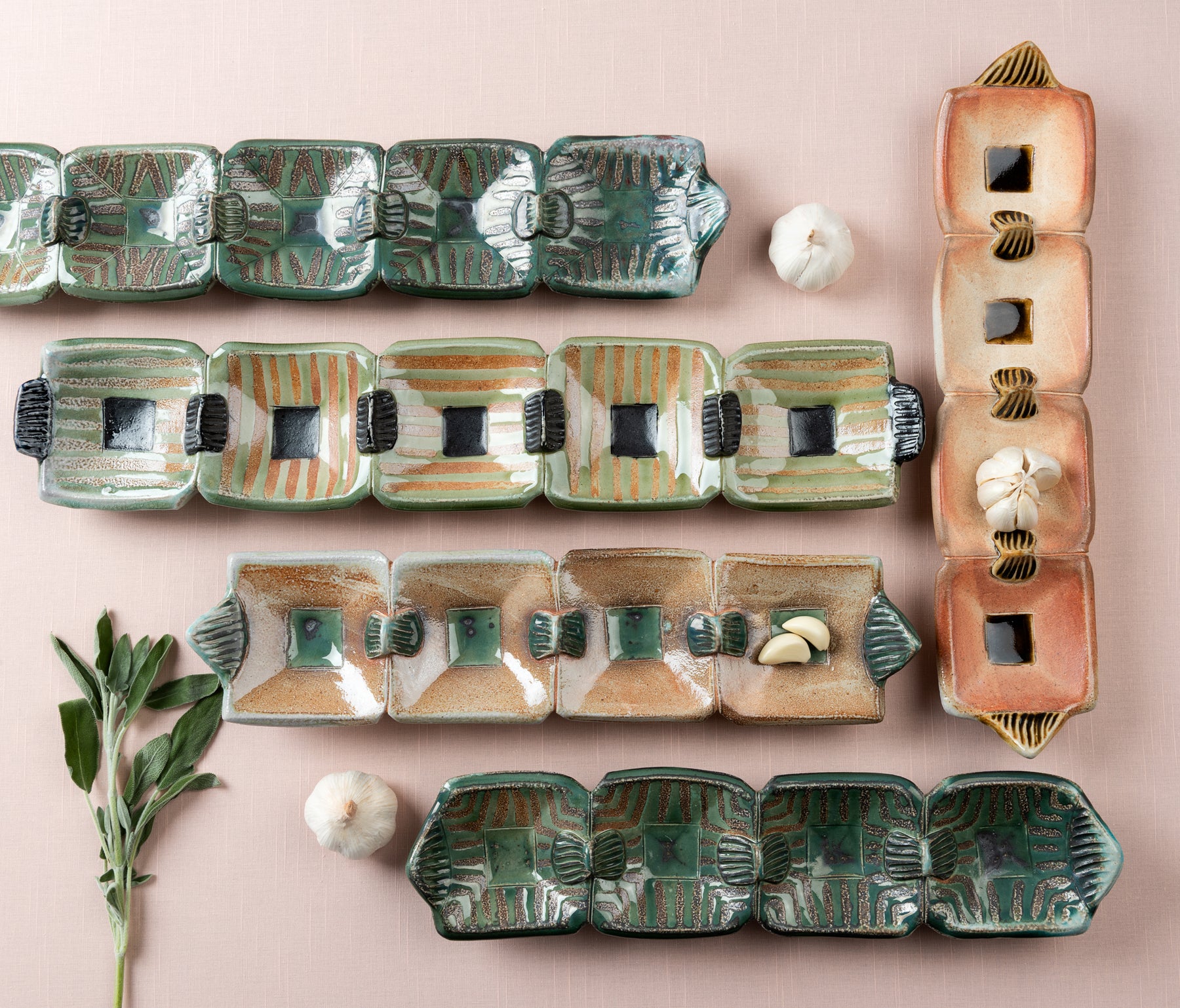 Five handmade and decorated rectangular snack trays, shot from above, sit on a pink table top with garlic and sage scattered about. Artist Joy Tanner. 