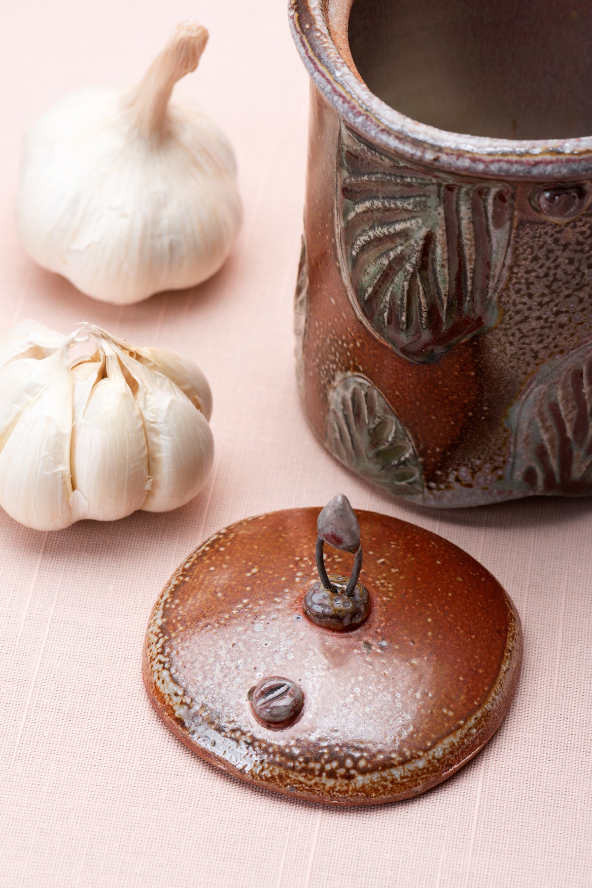 A detail of a handmade jar's lid on a peach colored tablecloth next to some heads of garlic. Artist Joy Tanner