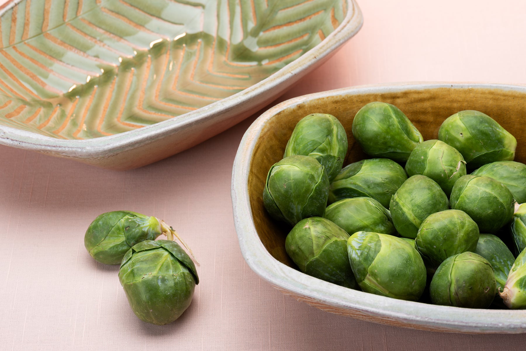Two rounded rectangular bowls sit on a pink table top. The pots are made by artist Joy Tanner and one of the bowls contains a little pile of brussels sprouts, and one is empty. 