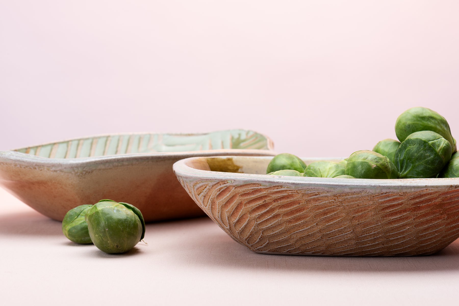 Two rounded rectangular bowls sit on a pink table top against a blush backdrop. The pots are made by artist Joy Tanner and one of the bowls contains a little pile of brussels sprouts, and one is empty.