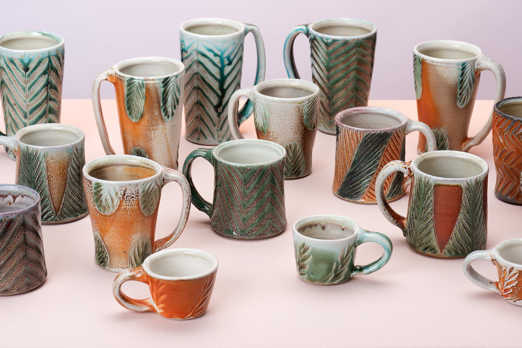 A group of different sized mugs decorated in different styles made by artist Joy Tanner sit on a peach table against a blush-purple  backdrop. Image by Loam 