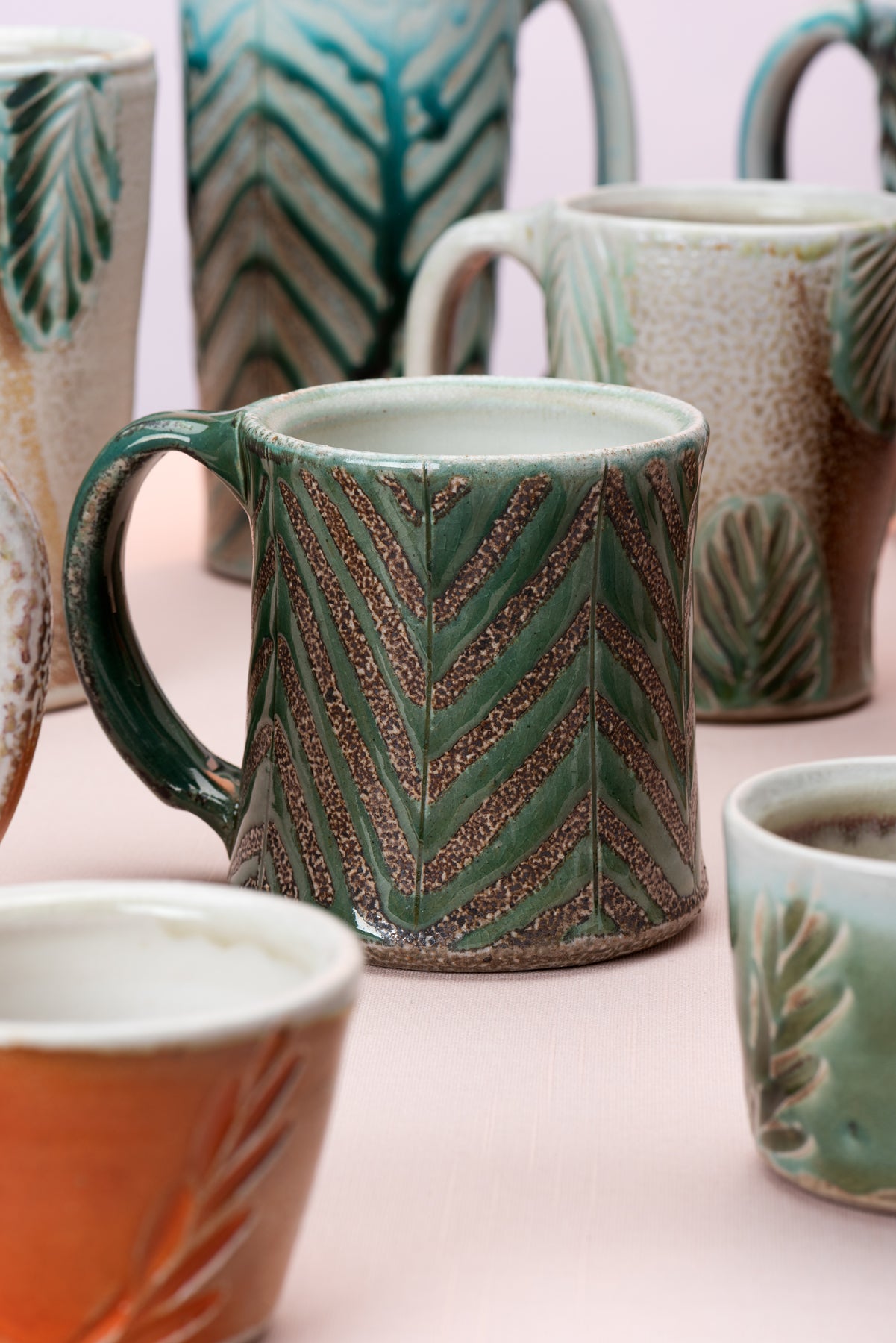 A detail of a group of mugs by potter Joy Tanner. One mug, featuring lines of turquoise and mottled brown, is in focus, while other mugs are out of focus in the foreground and background. 