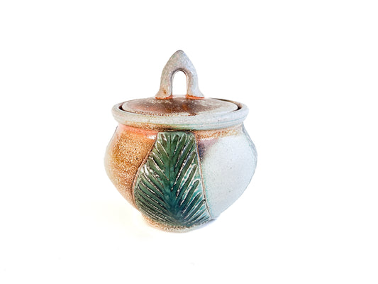 This small round handmade jar could be used to store tea bags, sugar, or candy. The green leaf pattern that is hand carved is inspired by nature. 