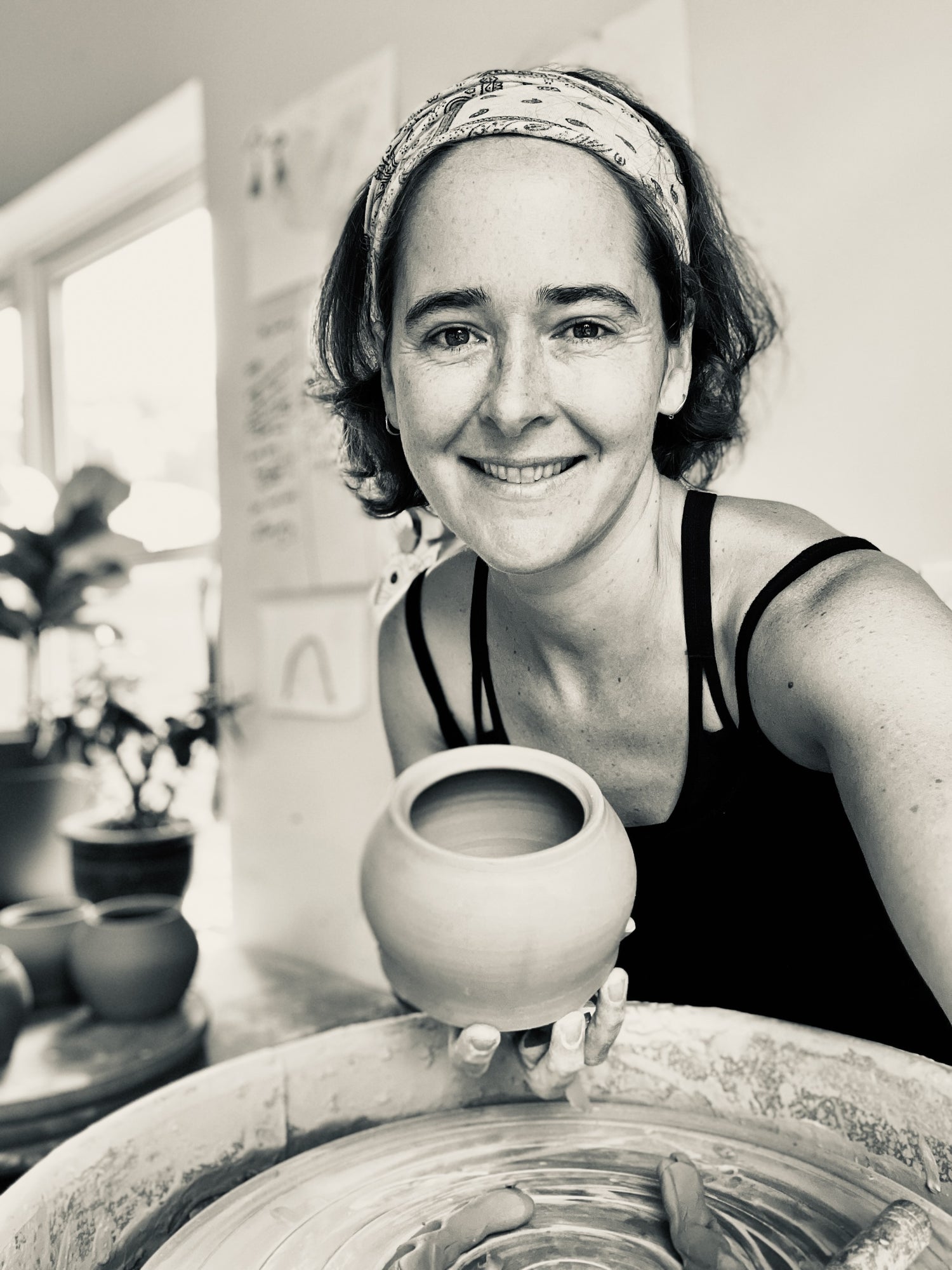 A black and white image of artist Joy Tanner sitting at her potters wheel, holding a pot in her hand and looking at the camera. 