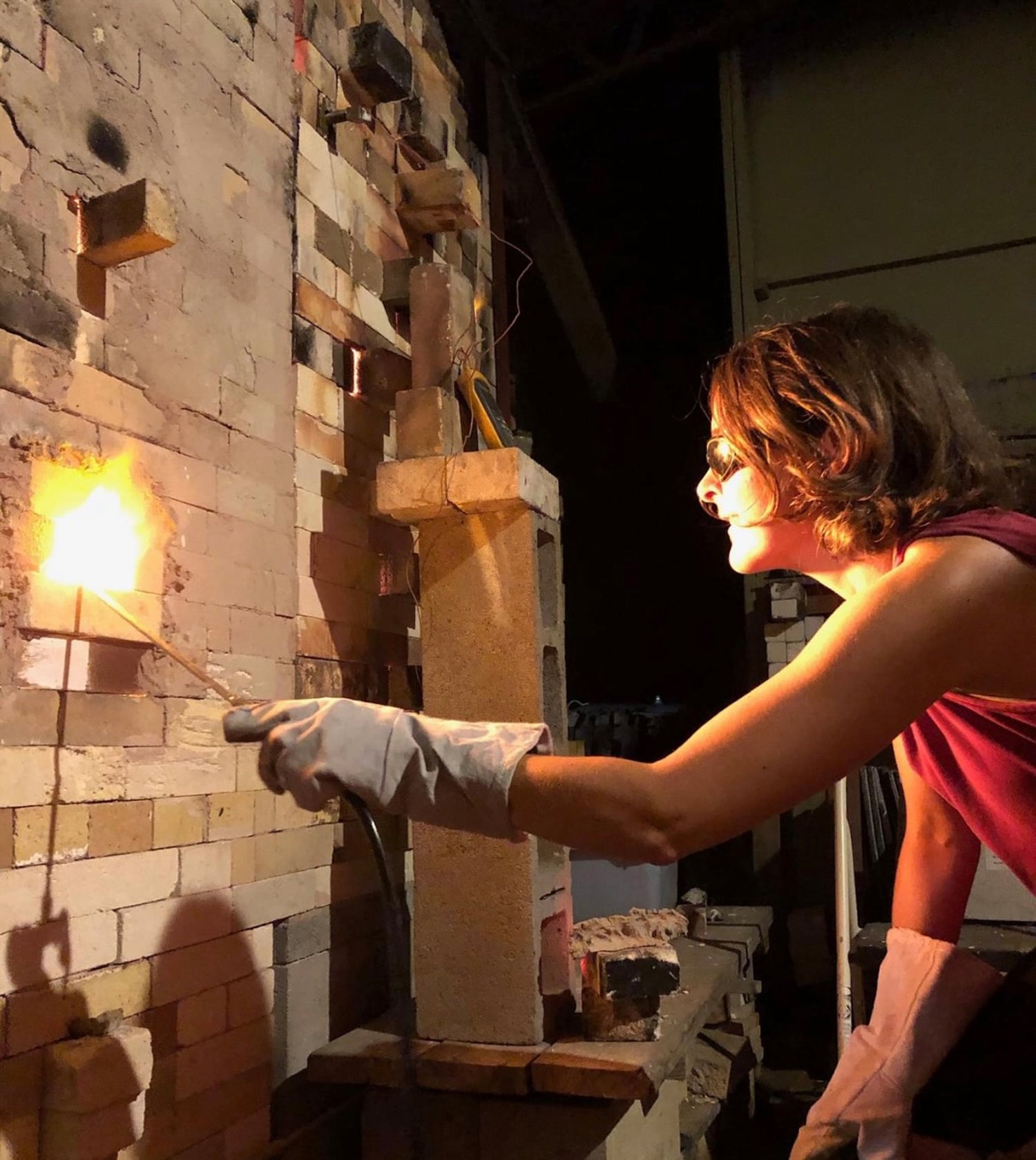 Joy Tanner pokes a rod into a white hot opening on the side of her kiln, the flames casting warm light on her face and sunglasses. 